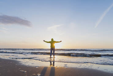 Full length rear view of senior man with arms outstretched standing on shore while looking at sea against sky during sunset, North Sea Coast, Flanders, Belgium - GWF06544