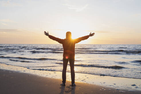 Full length rear view of woman with arms outstretched standing on shore while looking at sea against sky during sunset, North Sea Coast, Flanders, Belgium - GWF06545