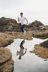 Männlicher Trailrunner in voller Länge, der auf Felsen läuft, mit seinem Spiegelbild über dem Wasser an der Küste, Ferrol, Galicien, Spanien - RAEF02371