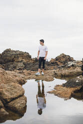 Männlicher Trailrunner in voller Länge auf Felsen stehend mit seinem Spiegelbild über dem Wasser gegen den Himmel, Ferrol, Galicien, Spanien - RAEF02372