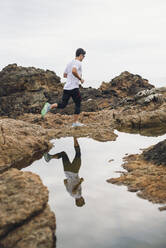 Männlicher Trailrunner in voller Länge auf Felsen, während er durch sein Spiegelbild über Wasser an der Küste läuft, Ferrol, Galicien, Spanien - RAEF02373