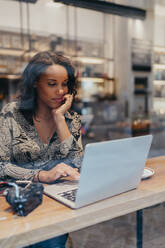 Young woman with a camera using laptop in a cafe behind windowpane - JPIF00462