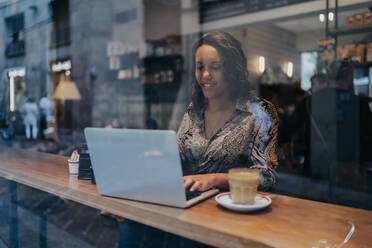 Young woman with a camera using laptop in a cafe behind windowpane - JPIF00463