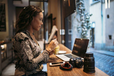 Junge Frau mit Kamera und Laptop in einem Cafe hinter einer Fensterscheibe - JPIF00466