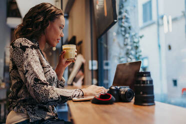 Junge Frau mit Kamera und Laptop in einem Cafe hinter einer Fensterscheibe - JPIF00467