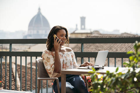 Junge Frau benutzt einen Laptop und telefoniert auf einem Balkon in Florenz, Italien, lizenzfreies Stockfoto
