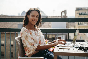 Portrait of smiling young woman using laptop on a balcony in Florence, Italy - JPIF00497