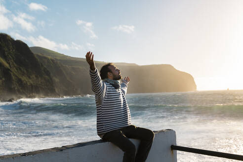 Man sitting at the coast at sunset with outstretched arms, Sao Miguel Island, Azores, Portugal - AFVF05697