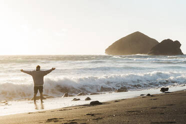 Mann steht bei Sonnenuntergang mit ausgestreckten Armen am Strand, Insel Sao Miguel, Azoren, Portugal - AFVF05705