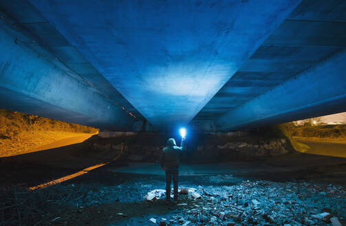Spanien, Galicien, Rückansicht eines vermummten Mannes unter einer Betonbrücke mit einem hellen blauen Licht in der Hand - RAEF02374