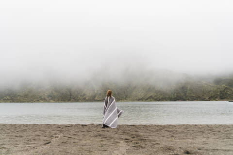 In eine Decke eingewickelte Frau am Seeufer auf der Insel Sao Miguel, Azoren, Portugal, lizenzfreies Stockfoto