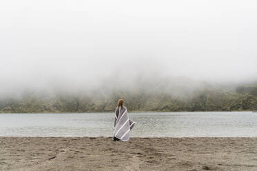 Woman wrapped in blanket standing at lakeshore in Sao Miguel Island, Azores, Portugal - AFVF05712
