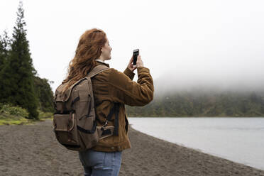 Weibliche Forscherin mit Rucksack beim Fotografieren mit dem Smartphone auf der Insel Sao Miguel, Azoren, Portugal - AFVF05719