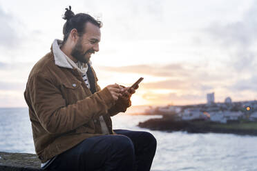 Lächelnder mittelgroßer erwachsener Mann, der ein Smartphone benutzt, während er am See auf der Insel Sao Miguel, Azoren, Portugal, sitzt - AFVF05721