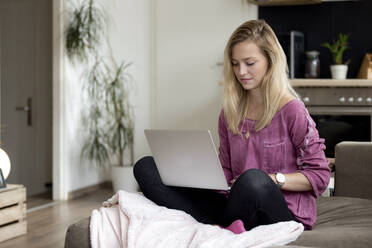 Portrait of blond young woman sitting on couch at home using laptop - FLLF00420
