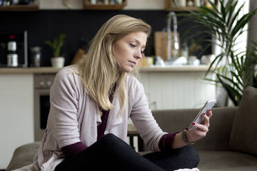 Porträt einer blonden jungen Frau, die zu Hause auf der Couch sitzt und auf ihr Mobiltelefon schaut - FLLF00421