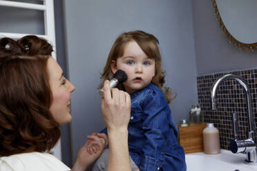 Smiling young mother applying rouge on her daughter's face in bathroom - AUF00135