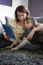 Junge Mutter und ihre kleine Tochter sitzen zu Hause auf dem Boden und sehen sich ein Bilderbuch an - AUF00142