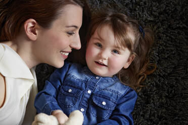 Portrait of happy toddler girl lying on carpet next to her mother - AUF00146