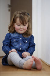 Portrait of smiling toddler girl sitting on the floor at home watching her bare feet - AUF00147