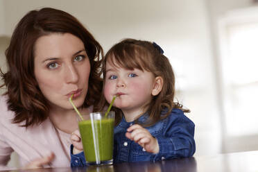 Porträt einer jungen Mutter und ihrer kleinen Tochter, die zusammen einen grünen Smoothie trinken - AUF00159