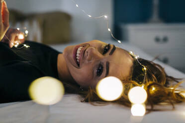 Porträt einer glücklichen jungen Frau im Bett liegend mit Lichterkette - DCRF00162