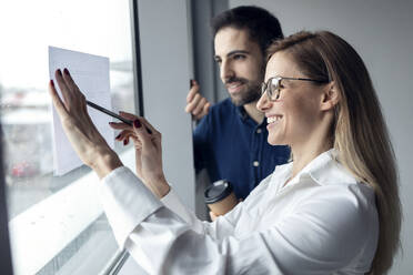 Businesswom holding sheet of paper on window, drawing chart with colleague watching - JSRF00898