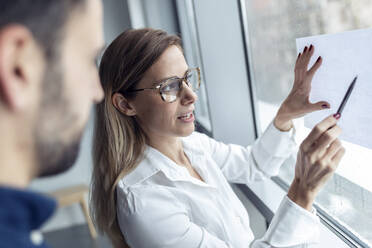 Businesswom holding sheet of paper on window, drawing chart with colleague watching - JSRF00900