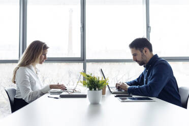 Businessman and woman sitting at desk, using laptops, looking seriously - JSRF00902