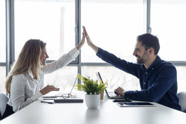 Businessman and woman sitting at desk, using laptops, high fiving - JSRF00904