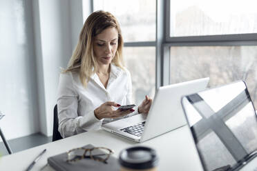 Businesswoman sitting in office working on laptop, using smartphone - JSRF00905