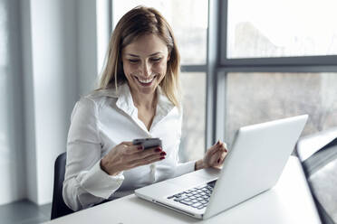 Businesswoman sitting in office working on laptop, using smartphone - JSRF00906