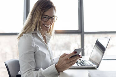 Businesswoman sitting in office working on laptop, using smartphone - JSRF00908