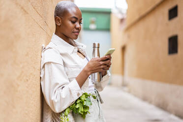 Woman leaning against a wall with thermos flask and groceries using smartphone - AFVF05752