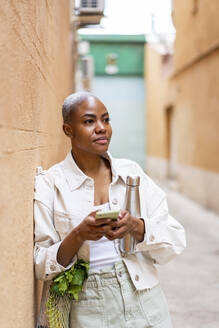 Woman leaning against a wall with thermos flask, groceries and smartphone - AFVF05753