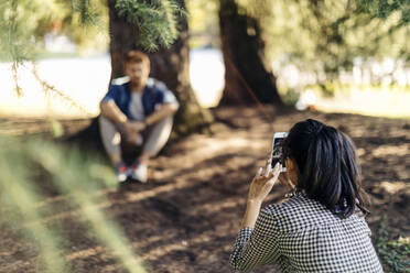 Young woman taking cell phone picture of boyfriend under a tree - SODF00737