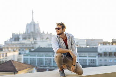 Young man crouching on a balcony above the city, Milan, Italy - SODF00746