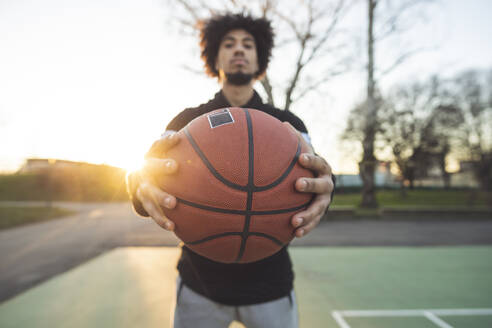 Junger sportlicher Mann posiert mit einem Basketball auf einem Platz bei Sonnenuntergang - MEUF00098