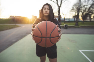 Junge sportliche Frau posiert mit einem Basketball auf einem Platz bei Sonnenuntergang - MEUF00099