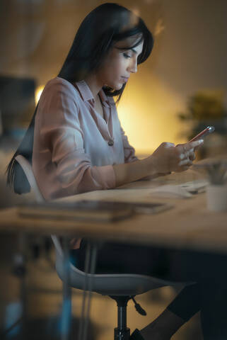 Junge Frau sitzt am Schreibtisch im Büro und schaut auf ihr Smartphone, lizenzfreies Stockfoto