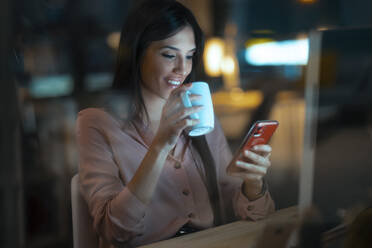 Lächelnde junge Frau mit Kaffeebecher sitzt am Schreibtisch im Büro und schaut auf ihr Smartphone - JSRF00922