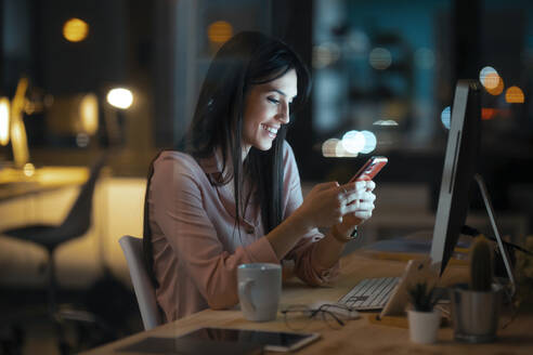 Smiling young woman sitting at desk in office looking at smartphone - JSRF00928