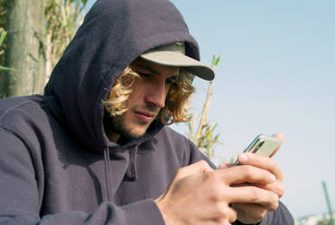 Nahaufnahme eines jungen Mannes mit Kapuze, der beim Entspannen am Strand von Tarifa, Spanien, sein Smartphone benutzt - OCMF01066