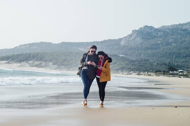 Volle Länge von glücklichen Freundinnen, die Mobiltelefone benutzen, während sie am Strand in Tarifa, Spanien, spazieren gehen - OCMF01068