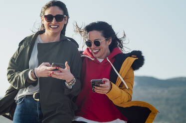 Fröhliche Freunde, die ein Mobiltelefon benutzen, während sie gemeinsam gegen den Himmel in Tarifa, Spanien, laufen - OCMF01069