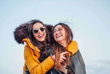 Fröhliche Frau, die ihre Freundin huckepack nimmt, während sie am Strand gegen den Himmel in Tarifa, Spanien, Spaß hat - OCMF01071