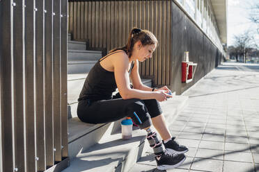 Sporty young woman with leg prosthesis sitting on stairs in the city using smartphone - MEUF00112