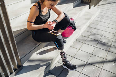 Sportliche junge Frau mit Beinprothese sitzt auf einer Treppe in der Stadt und benutzt eine Smartwatch - MEUF00113
