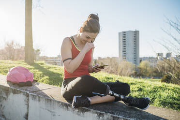 Sportliche junge Frau mit Beinprothese, die auf einer Mauer sitzt und ein Mobiltelefon benutzt - MEUF00154