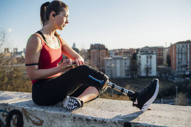 Sporty young woman with leg prosthesis sitting on a wall above the city, Milan, Italy - MEUF00162